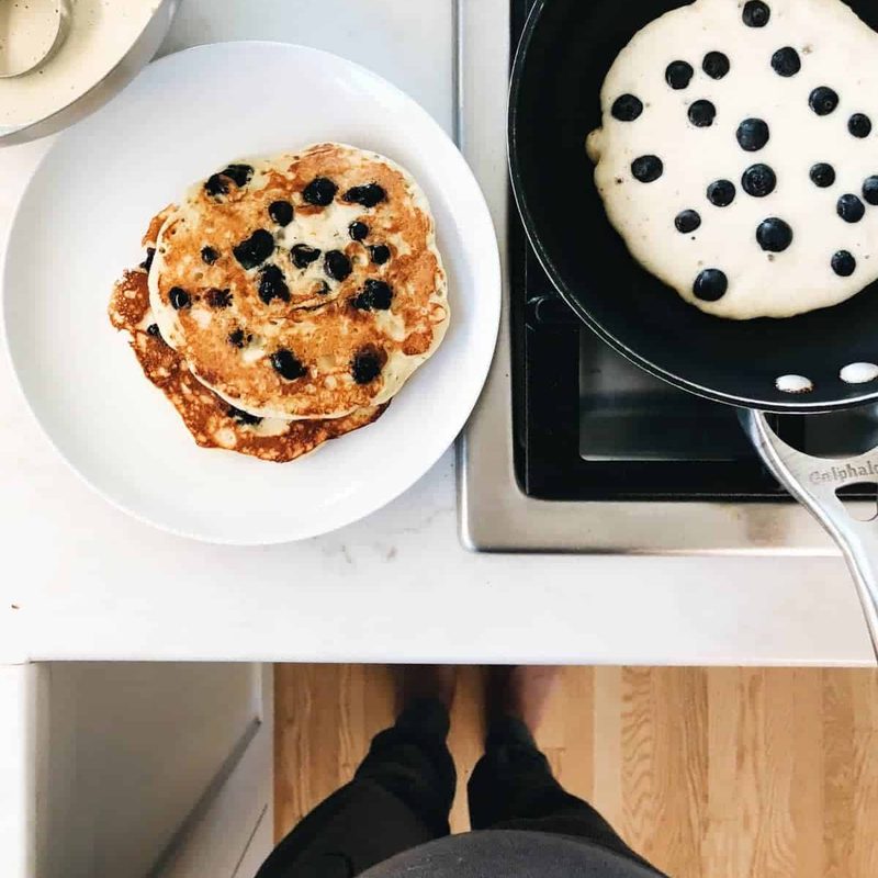 A large white plate of blueberry pancakes with butter melting over them as some cook in a skillet next to it.